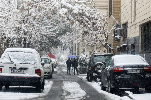 Streets blanketed in heavy snow as residents navigate through a snow-covered neighborhood under an umbrella, highlighting the winter conditions impacting daily life in Iran.