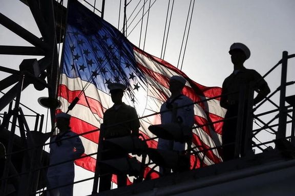A US flag flutters in the background as members of the Marines and the Navy stand on a deck of the USS Bataan, a Navy Wasp-class amphibious assault ship, deployed to the Persian Gulf in July.