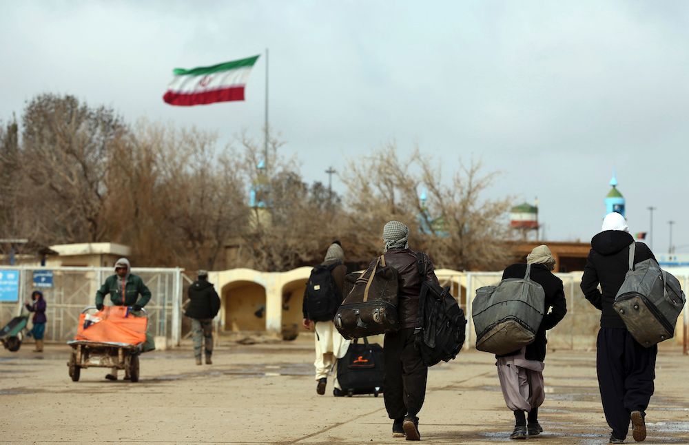 Afghan migrants walk away with their belongings as the Iranian flag flies in the background