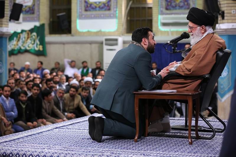 A maddah kneeling on front of Khamenei tokiss his hand