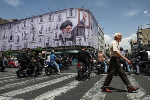 A man crosses a street in Tehran as motorcyclists pass beneath a giant billboard depicting Iran’s slain supreme leader Ali Khamenei, May 6, 2026