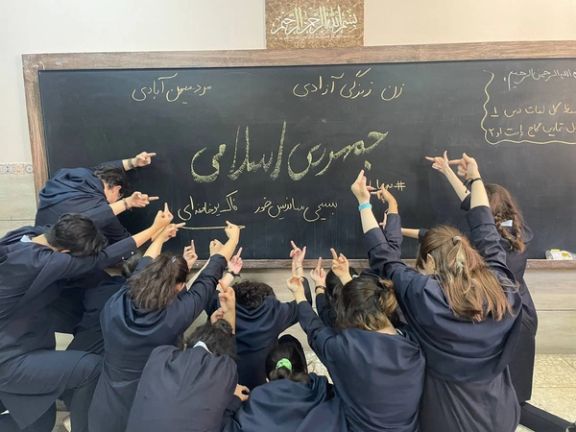 Iranian schoolgirls showing finger to a blackboard with “the Islamic Republic” words written on it
