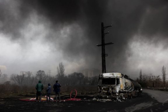 People stand near a destroyed vehicle as smoke rises after a reported strike on Shahran fuel tanks, amid the US-Israeli conflict with Iran, as seen through a window, in Tehran, Iran, March 8, 2026.